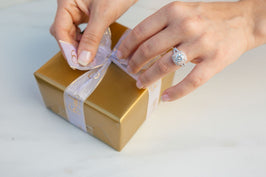 person tying white bow onto gold gift box with diamond ring on finger