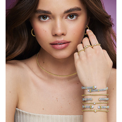 Woman wearing gold jewelry including earrings, necklace, and bracelets on a plain background.